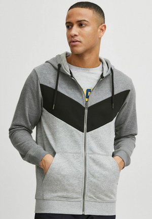 Siva zip-up kapuca z črnim chevron vzorcem, nastavljiva kapuca, sprednji žepi in teksturirana tkanina. Ima rebraste manšete in rob.