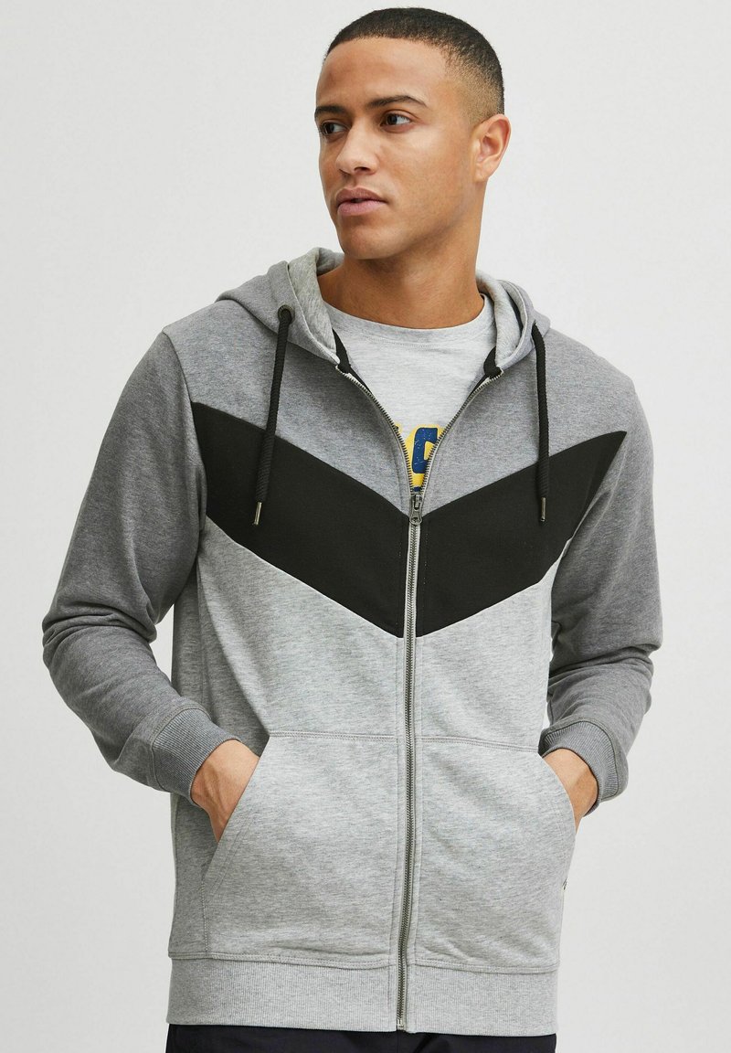 Gray zip-up hoodie with black chevron design, adjustable hood, front pockets, and textured fabric. Features ribbed cuffs and hem.