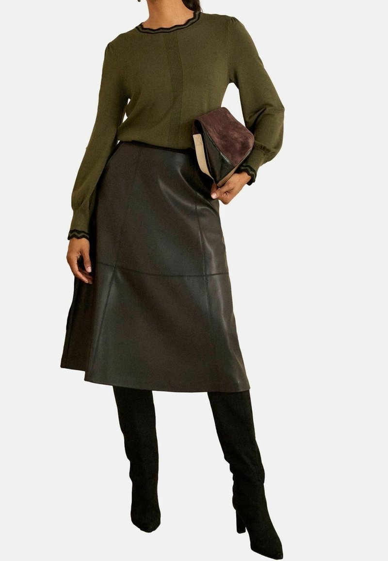Femme portant un pull à manches longues vert olive, une jupe midi en cuir marron foncé, des bottes noires montantes jusqu'aux genoux, tenant une pochette marron et beige.