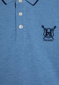 Svetlomodrá polo tričko vyrobené z textúrovanej tkaniny, s tromi bielymi gombíkmi a tmavomodrým vyšitým logom "Hackett" s prekríženými veslami.