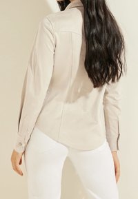 Camisa de manga larga color beige claro con textura suave, puños abotonados y silueta ajustada, combinada con pantalones blancos.