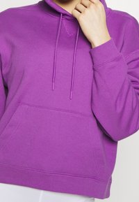 Felpa viola con tasca a marsupio, lacci e polsini a costine. Il tessuto sembra morbido con una texture liscia e una vestibilità comoda.