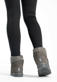 Botas de ante grises hasta el tobillo con puños de punto y suelas texturizadas, combinadas con pantalones negros ajustados, vistas desde atrás.