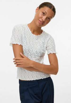 Donna con capelli intrecciati, indossante una maglietta bianca in pizzo a maniche corte e pantaloni blu navy, in posa con le braccia incrociate contro uno sfondo bianco.