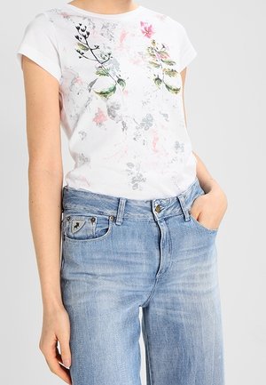Camiseta blanca con estampado floral en verde, rosa y gris; de manga corta. Combinada con jeans de tiro alto en azul claro con un acento de logo.
