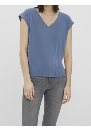 Kobieta w niebieskiej bluzce z krótkim rękawem i dekoltem w serek oraz szarych przylegających dżinsach na jednolitym jasnym tle.