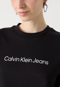 Čierne bavlnené tričko s okrúhlym výstrihom má na prednej strane umiestnený biely grafický nápis "Calvin Klein Jeans".
