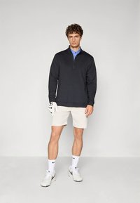 Sweatshirt preta com fecho zip sobre uma polo azul, combinada com calções bege e sapatilhas brancas. O modelo usa uma luva de golfe numa mão.