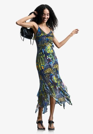 Kobieta w kolorowej, tropikalnej sukience bez rękawów, trzymająca czarną torebkę na ramieniu, ubrana w czarne sandały, uśmiechająca się.