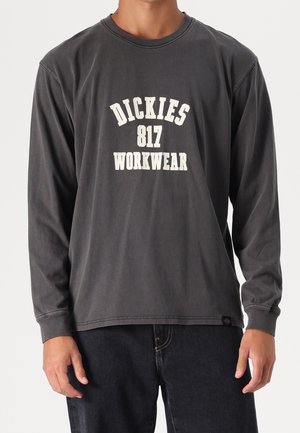 Henkilö, joka käyttää haalistunutta mustaa pitkähihnaista paitaa, jossa on valkoinen teksti "DICKIES 817 WORKWEAR", sekä mustia housuja.