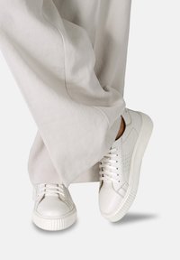 Baskets en cuir blanc avec des détails texturés, semelles en caoutchouc plates et orteils ronds, assorties à un pantalon en tissu gris clair ample.