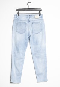 Světle modré džíny z denimu visí na černém ramínku, mají pět kapes, zadní nášivku a mírně vybledlou texturu.