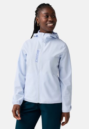 Femme portant une veste zippée bleu clair avec le logo "BROOKS", un pantalon foncé, souriante et regardant vers sa gauche sur un fond uni.