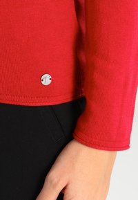 Czerwony sweter z dzianiny z okrągłą srebrną metką u dołu, prezentujący gładką fakturę i dopasowany krój, w połączeniu z czarnymi spodniami.