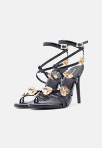 Μαύρα σανδάλια ψηλοτάκουνα από λείο δέρμα με πολλές διασταυρούμενες λουρίδες. Το μπροστινό μέρος διακοσμημένο με χρυσές ανθικές λεπτομέρειες. Λείο υλικό και μυτερή μύτη.