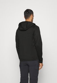 Man som bär svart hooded jacka och mörka cargobyxor, vänd bort från enkel bakgrund.