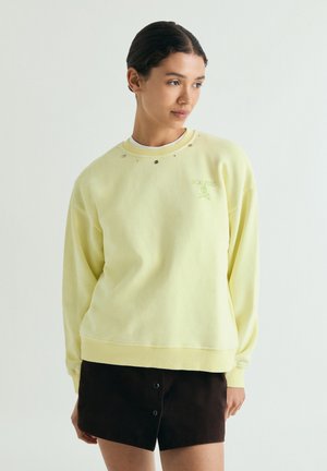 Kvinde med mørkt hår iført en sart gul sweatshirt og sort nederdel, der kigger lidt til siden mod en ensartet lys baggrund.