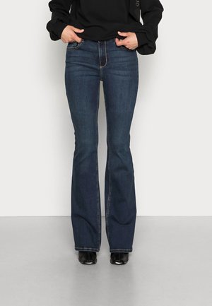 Person iført mørkeblå vidde jeans, sorte ankelstøvler og en sort langærmet top med hænder, der justerer jeansens taljebånd.