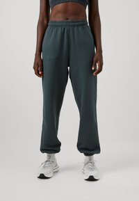 Sötétzöld jogger nadrág, puha anyagból, rugalmas derékkal és szűkített szárakkal, világosszürke sportcipőkkel párosítva.