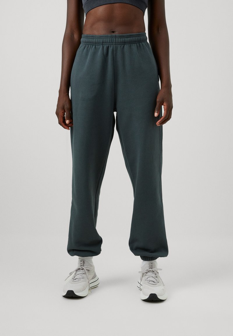 Sötétzöld jogger nadrág, puha anyagból, rugalmas derékkal és szűkített szárakkal, világosszürke sportcipőkkel párosítva.