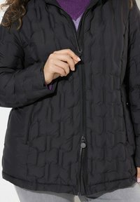 Svart vadderad jacka med dragkedja fram, sidofickor och ett texturerat tyg med en slät finish. Bärs över en lila tröja.