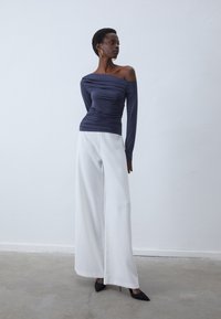 Marinblus med off-shoulder-design och rynkdetaljer, matchad med vita vida byxor och svarta spetsiga klackar, mot en ljus bakgrund.