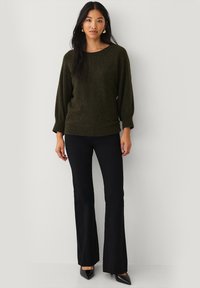 Pull en maille vert olive avec un décolleté ample et des manches 3/4, associé à un pantalon noir évasé et des chaussures noires à bout pointu.