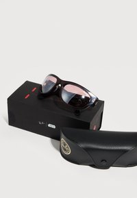 Γυαλιά ηλίου Ray-Ban με σκούρο σκελετό και φακούς σε ροζ διαβάθμιση, τοποθετημένα πάνω σε μαύρο κουτί με την επιγραφή "Ray-Ban" και "Star Wars", δίπλα σε μία μαύρη θήκη.