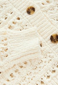 Cardigan en crème tricoté avec un motif texturé, des bords côtelés et deux boutons ronds marron. Gros plan sur le détail de la manche et du corps.