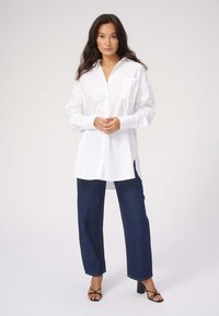 Hvid skjorte med knapper og en brystlomme, lange ærmer og en afslappet pasform, parret med mørkeblå vide bukser og sorte hæle sandaler.