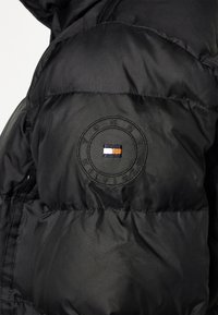 Doudoune noire avec un logo brodé circulaire portant le texte "TOMMY HILFIGER" et un détail de rayure horizontale multicolore.