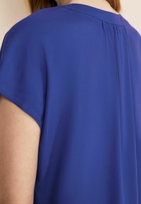 Královsky modrá blůza s krátkým rukávem; lehký, hladký materiál; jemný detail v zadní části v podobě sklady. Volný střih, kulatý výstřih, bez viditelných zapínání.
