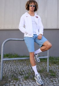 Valkoinen kevyt takki, jossa on punainen logo, istuva tanktoppi, siniset farkkushortsit, valkoiset nilkkasukat ja harmaat lenkkarit. Ulkotila, jossa on soraa.