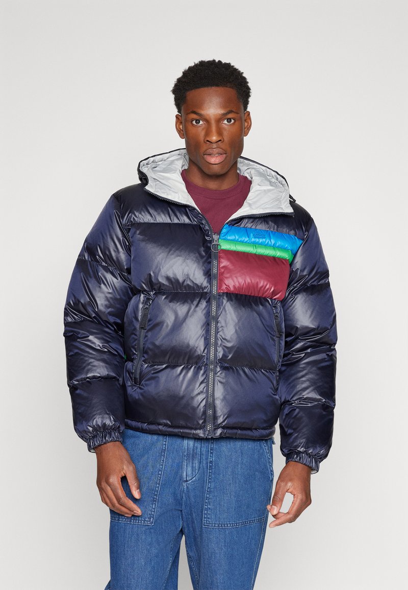 Marineblaue Pufferjacke mit grauem Kapuzenbereich, die ein mehrfarbiges Brustpanel in Rot, Grün und Blau aufweist. Mit Reißverschluss versehene Fronttaschen und elastische Bündchen.