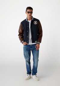 Chaqueta varsity navy con mangas de cuero marrón, puños con rayas blancas y un parche en el pecho, combinada con jeans de mezclilla azul y zapatillas blancas.