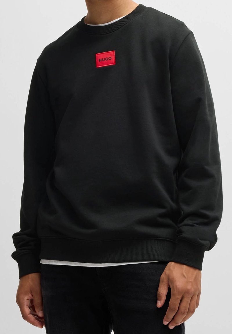 Sweat-shirt noir à manches longues et col rond avec un patch rectangulaire rouge affichant "HUGO" sur la poitrine, porté sur un sous-vêtement blanc.