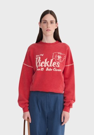 Femme portant un sweat-shirt rouge avec le texte "Pickles" et un motif de chien, assorti à un pantalon bleu, tenant un sac à bandoulière marron sur un fond blanc.