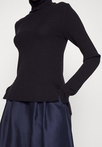 Pull-over noir à col roulé côtelé avec manches longues et poches latérales, présentant une silhouette ajustée et un motif en tricot texturé.
