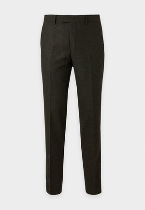 Homme portant un polo tricoté beige clair à manches courtes, pantalon foncé et chaussures noires à lacets, debout les mains dans les poches.