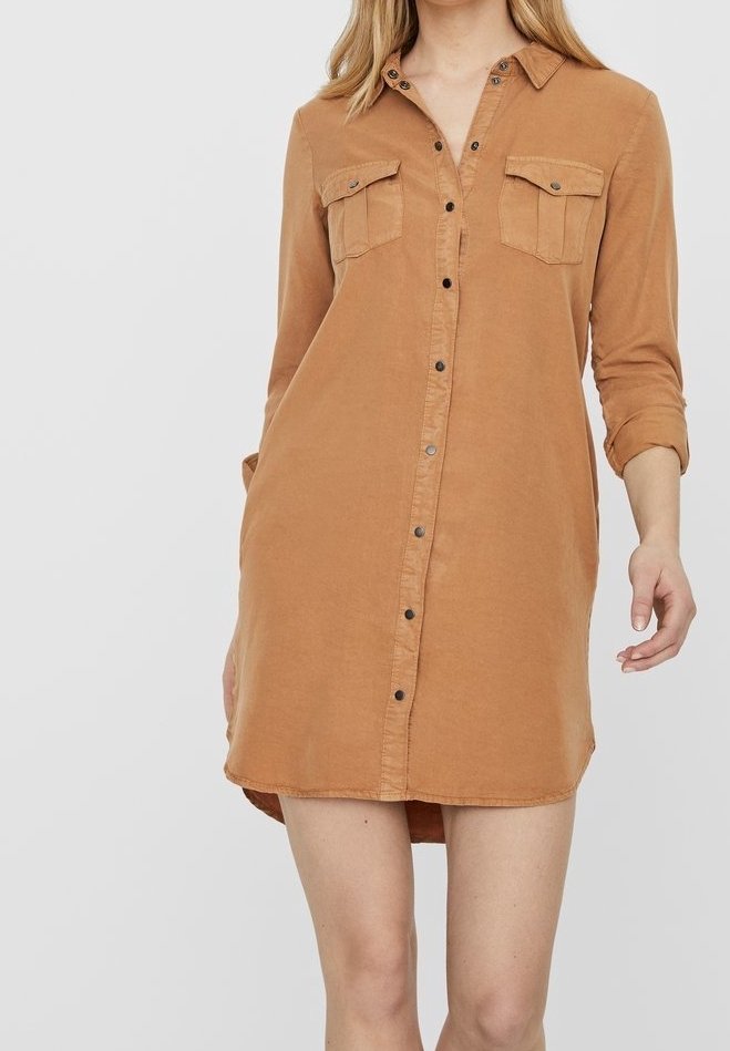 Femme portant une robe chemise marron clair boutonnée, avec des manches longues retroussées, dotée de deux poches poitrine et un col.