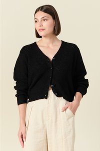 Cardigan en tricot noir avec un col en V, six boutons, des poignets côtelés et une texture géométrique complexe. Assorti à un pantalon beige clair.