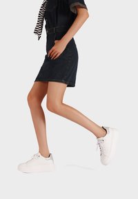 Donna in un vestito di denim scuro e sneaker bianche con piattaforma che cammina su uno sfondo grigio chiaro uniforme.