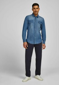 Denimhemd in Mittelblau mit klassischem Kragen, zwei Brusttaschen und Knöpfen auf der Vorderseite. Kombiniert mit dunklen Hosen und weißen Sneakers.