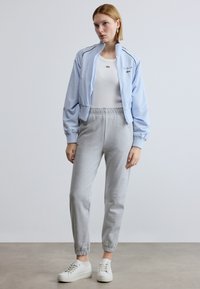 Gaiši zila bombers jaka ar rāvējslēdzēju, balta pieguloša t-krekls, pelēkas sporta bikses ar aprocēm un balti sporta apavi. Lacoste logotips uz jakas.