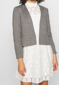 Blazer gris court en tissu texturé, avec un devant ouvert et des lignes épurées. Porté sur une robe blanche en dentelle avec des motifs floraux.