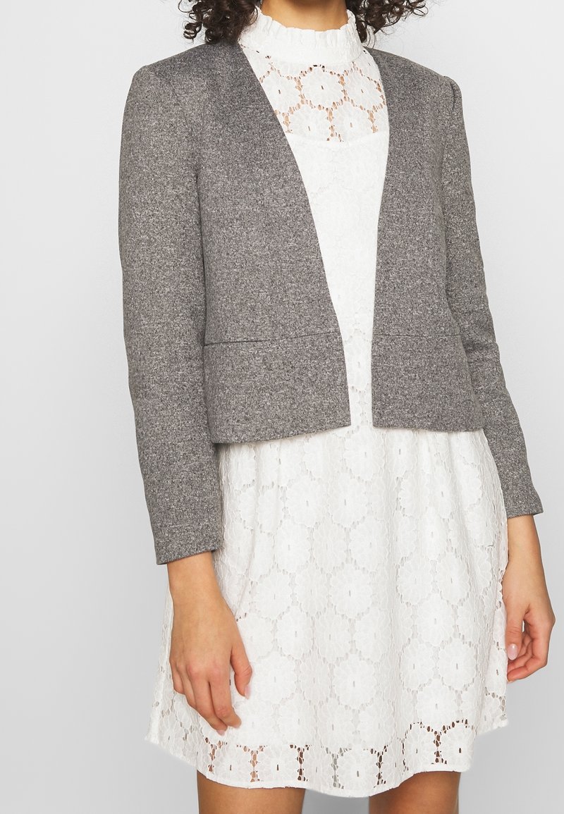 Blazer gris court en tissu texturé, avec un devant ouvert et des lignes épurées. Porté sur une robe blanche en dentelle avec des motifs floraux.
