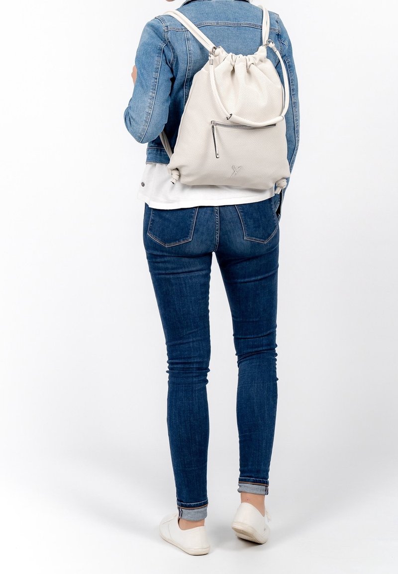 Sac à dos couleur crème avec fermeture à cordon, surface texturée, poche zippée à l'avant et bretelles ajustables, porté sur le dos.