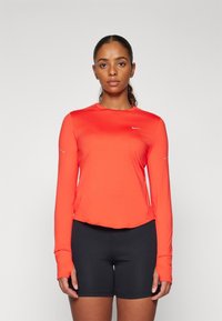 Langärmliges Sportshirt aus glattem, leuchtend rotem Stoff mit abgerundetem Saum und kleinem weißen Logo auf der Brust, kombiniert mit schwarzen Shorts.