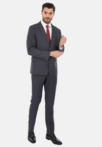 Completo slim-fit grigio antracite con rever a punta, chiusura a un bottone; abbinato a una camicia bianca e cravatta rossa; scarpe nere lucide.
