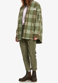Zelená kostkovaná nadměrná bunda s vysokým límcem, zapínáním na zip a žebrovaným lemem, doplněná olivovými kordurovými kalhotami a hnědými kotníkovými botami.
